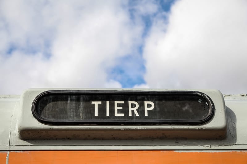 Bilden visar en detaljbild av en ljusgrå skylt med svart textfält där ordet "TIERP" står skrivet i vita versaler. Skylten är inramad med en svart kant. Under den ljusgrå skylten syns en orange yta. I bakgrunden syns en blå himmel med vita moln som sk