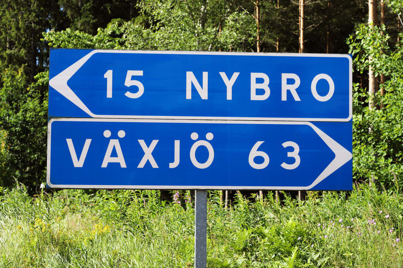 En blå vägskylt med vit text är placerad utomhus, delvis omgiven av grön vegetation och träd, som tallar och lövträd. Övre delen av skylten visar '15 NYBRO' med en vänsterpekande pil. Nedre delen visar 'VÄXJÖ 63' med en högerpekande pil. Texten är vi
