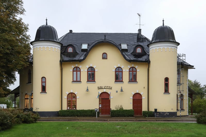 Bilden visar fasaden på en gul byggnad med en svart kupolformad takkonstruktion och mörkgrå takpannor. Byggnaden har två framträdande runda torn med svarta kupoler, ett på vardera sida. Mellan tornen finns sex fönster med röda karmar och flera takkup