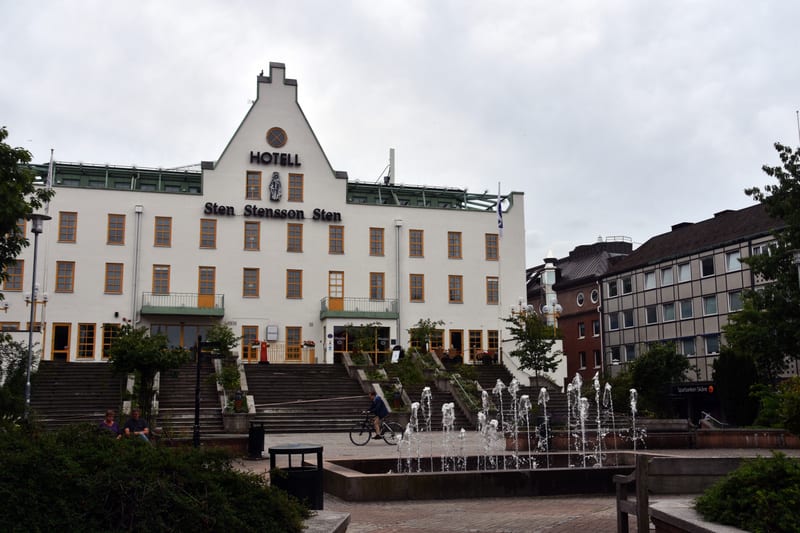 Bilden visar ett torg med en stor vit byggnad i bakgrunden som har texten "HOTELL Sten Stensson Sten" på fasaden. Byggnaden har många fönster med gula ramar och flera entréer med trappor som leder upp till dem. Framför hotellet finns en bred trappa s