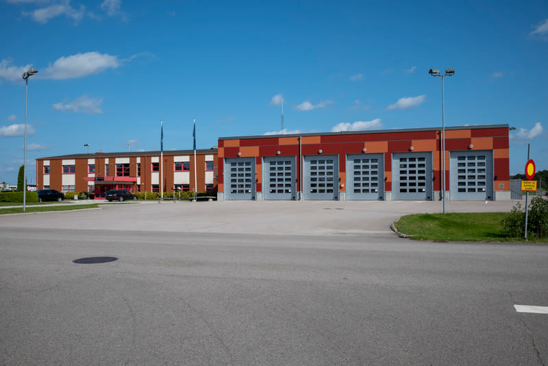 En exteriörbild som visar en stor byggnad bestående av två sammanlänkade delar under en klarblå himmel med några vita moln. Den vänstra delen av byggnaden är i huvudsak brun med röda detaljer runt fönster och dörrar. Framför denna del syns ett antal