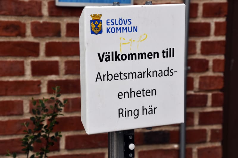 En vit skylt med svart text och en logotyp är framträdande i bilden. Skylten bär texten "ESLÖVS KOMMUN" under en logotyp med ett vapen. Under kommunnamnet står "Välkommen till Arbetsmarknads-enheten Ring här". Ovanför "Välkommen till" har någon klott