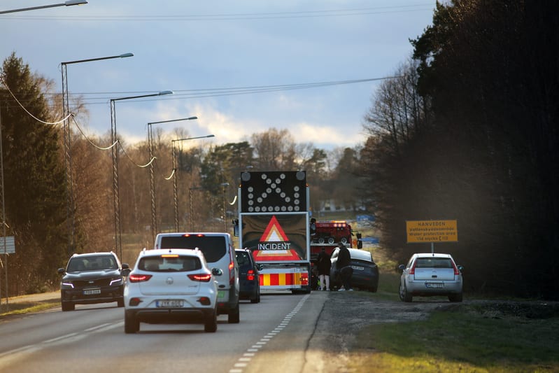 Bilden visar en trafiksituation på en väg under dagtid. Flera bilar är synliga på vägen, varav några står stilla i vad som ser ut som en kö eller en incident. Längre fram på vägen står ett fordon med en stor digital skylt som visar en pil som pekar å
