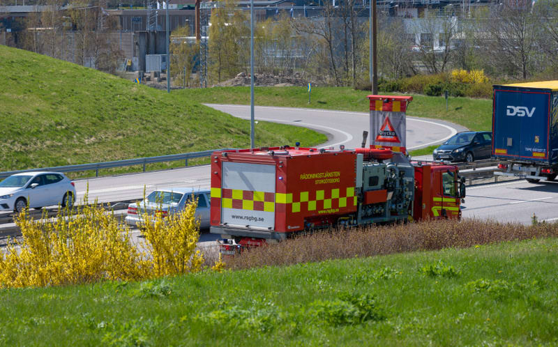 Bilden visar en vägmiljö under dagtid. I förgrunden syns en grässlänt med gula blommande buskar, sannolikt forsythia. På vägen, som har flera filer, står ett rött räddningsfordon med texten "RÄDDNINGSTJÄNSTEN STOR-GÖTEBORG" och en webbadress "www.rsg