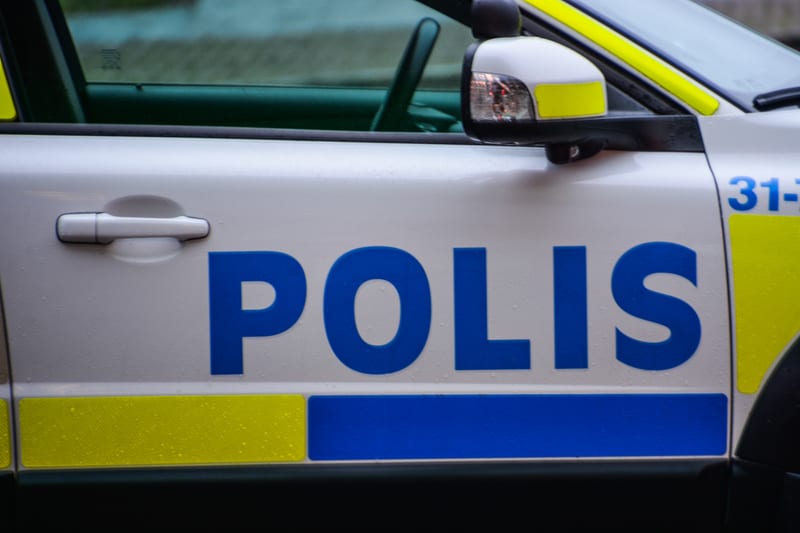 Bilden visar närbilder på den högra sidan av en svensk polisbil. Huvudfokus ligger på mitten av dörren, där det blåa ordet "POLIS" syns tydligt mot den vita bakgrunden, med gult och blått reflekterande material under texten. Det finns vattendroppar p