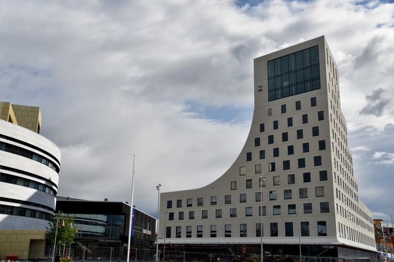 Bilden visar flera moderna byggnader under en delvis molnig himmel. I förgrunden till höger dominerar en hög, ljus byggnad med en distinkt, svängd form. Den har många kvadratiska fönster som är ojämnt fördelade över fasaden. Överst på denna byggnad f