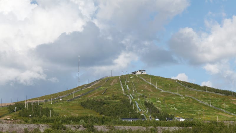 Bilden visar en bergssluttning täckt av grön vegetation och en del buskar under en mestadels molnig himmel. Sluttningen är utrustad med flera skidliftar eller liknande anläggningar, bestående av stolpar med ledningar och sittplatser eller byglar. Det