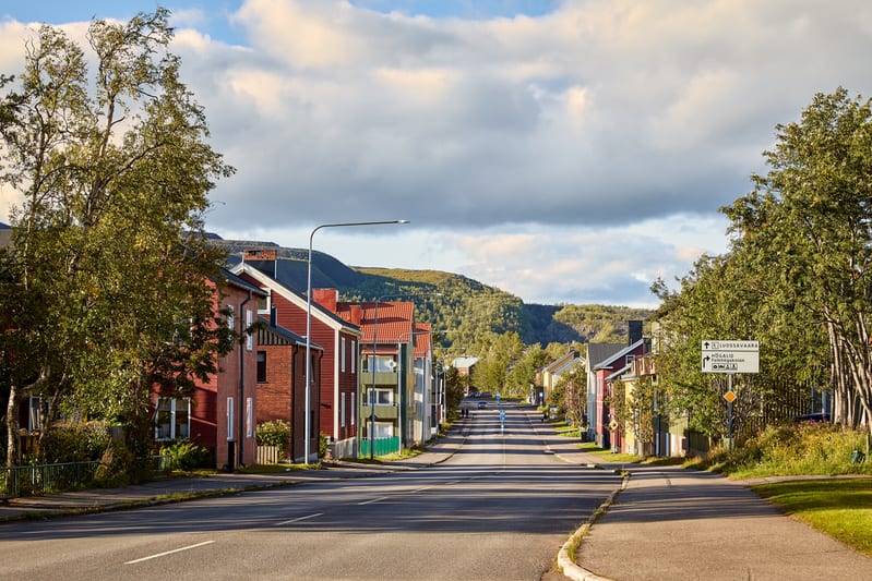 En gata i en stad med hus på båda sidor, under en delvis molnig himmel. Husen på vänster sida av gatan är mestadels röda och tegelröda, med några mindre byggnader och träd framför. På höger sida av gatan syns en blandning av hus i olika färger, samt