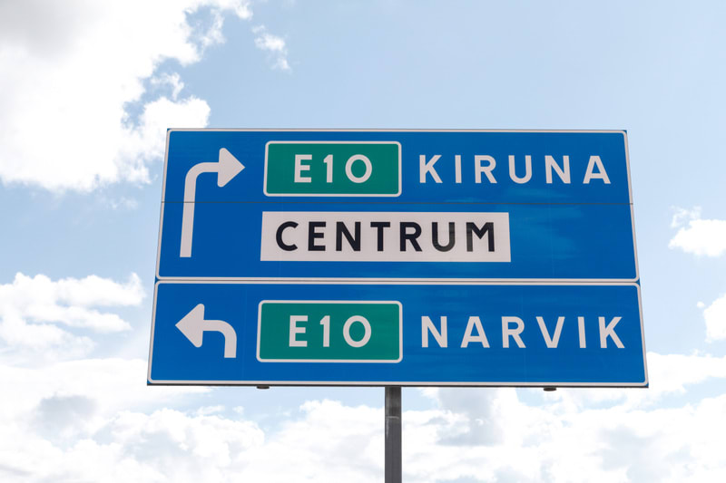 Bilden visar en blå vägskylt med vit text och gröna fält, monterad på en grå stolpe under en ljusblå himmel med spridda vita moln. Skylten är uppdelad i två sektioner. Den övre sektionen ger anvisningar mot E10 Kiruna Centrum, där "Kiruna" och "Centr