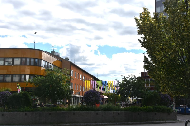 Bilden visar ett torg eller en öppen plats omgiven av byggnader i stadsmiljö under dagtid. I förgrunden syns en låg stenmur eller kant som avgränsar ett område med grönområden och buskar. Flera träd med gröna löv är utspridda över ytan. I mitten av b