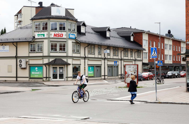 Bilden visar en gatuscen med flera byggnader och personer. I förgrunden syns en person som cyklar på en blå cykel, samt en person som korsar gatan vid ett övergångsställe. Byggnaderna i bakgrunden har olika fasader, bland annat en ljus byggnad med mö