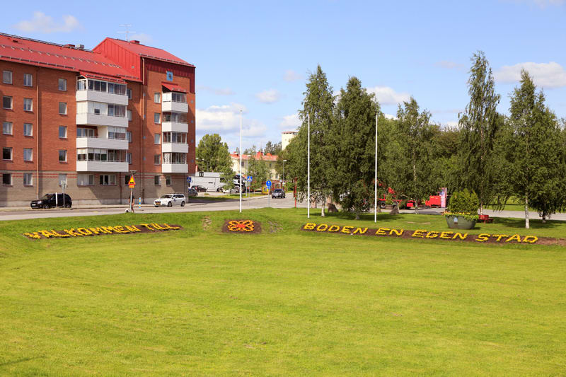 Bilden visar en utomhusmiljö med en gräsmatta i förgrunden, där texten "VÄLKOMMEN TILL" och "BODEN EN EGEN STAD" är formad med gula blommor. Mellan textraderna finns en mindre, rund formad med röd-orange blommor. I bakgrunden till vänster står ett rö