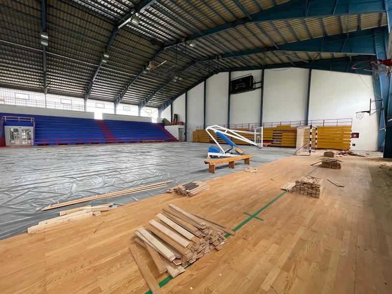 Bilden visar insidan av en stor idrottshall som är under renovering eller underhåll. En del av golvet är täckt med en grå presenning, medan en annan del består av ett ljust trägolv. På trägolvet ligger högar med träplankor, troligen för golvläggning.