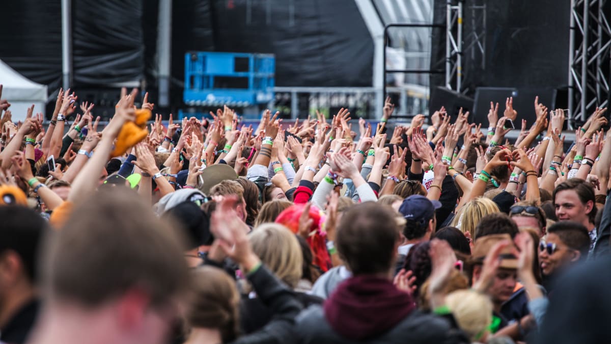 Bilden visar en folkmassa framför en scen utomhus, troligen under ett evenemang eller en konsert. Många händer är uppsträckta i luften med olika gester, som till exempel Peace-tecknet (V-form), knutna nävar eller öppna handflator. Flera personer bär