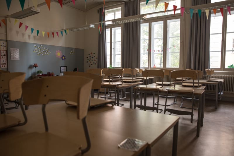 En bild inifrån ett klassrum med flera trästolar och bord. Stolar är resta upp på borden, vilket tyder på att klassrummet är tomt eller stängd för dagen. Väggen i bakgrunden är dekorerad med färgglada pappersdekorationer, inklusive flaggirlanger och
