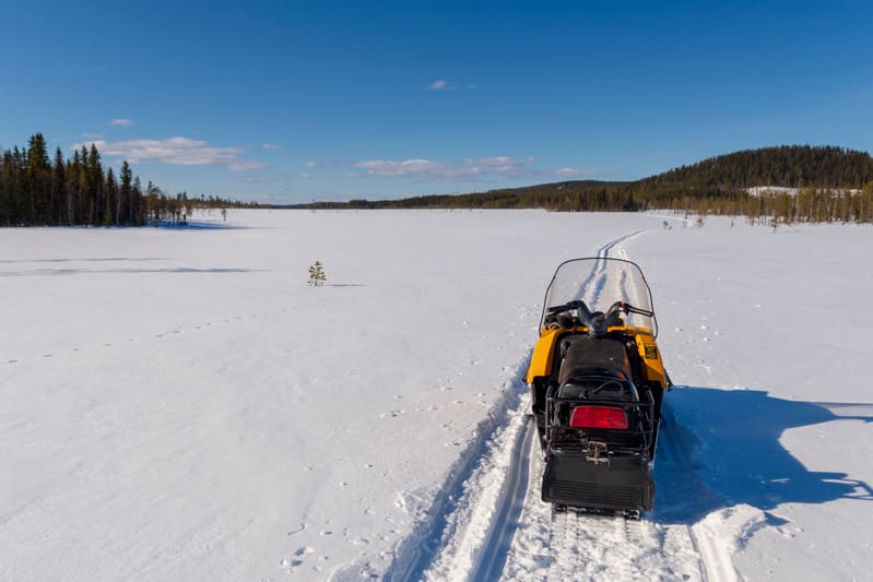 På bilden syns en gul snöskoter parkerad i snön. Framför snöskotern sträcker sig ett uppkört spår genom det snötäckta landskapet. Ovanför horisonten syns en klarblå himmel med några vita moln. I bakgrunden syns snötäckta fält som sträcker sig mot en