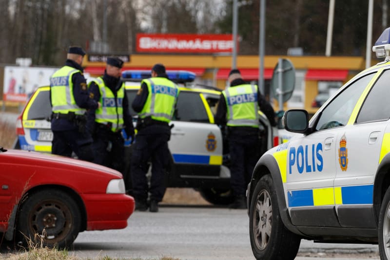 Bilden visar flera poliser och polisbilar utomhus under dagtid. I förgrunden till höger syns en del av en svensk polisbil i vitt med blå och gula ränder samt texten "POLIS" i blått. Till vänster i förgrunden syns fronten av en röd personbil, delvis b