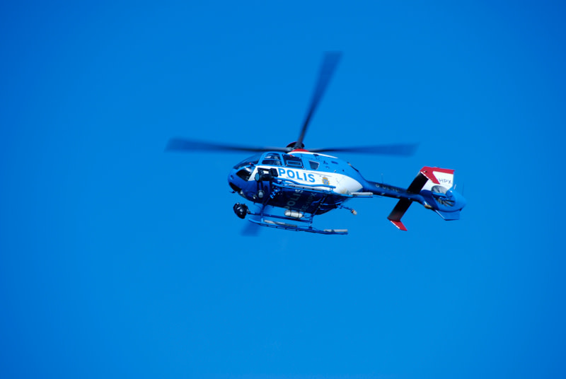 Bilden visar en polisflykopter i luften mot en klarblå himmel. Helikoptern är blå och vit med texten "POLIS" tydligt synlig på sidan. Rotorbladen är i rörelse, vilket indikeras av viss oskärpa. En del av helikopterns stjärtparti, med röda och vita de