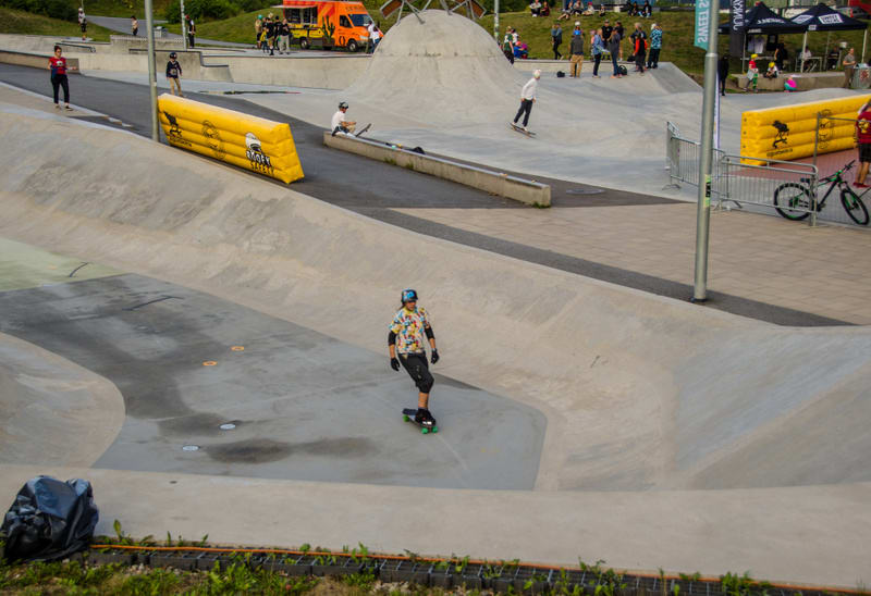 Bilden visar en utomhus skatepark med flera personer. I förgrunden åker en person skateboard på en av betongramperna, iklädd hjälm, en färgglad tröja och svarta skydd. I bakgrunden syns fler betongstrukturer, inklusive en stor kulle och andra ramper.