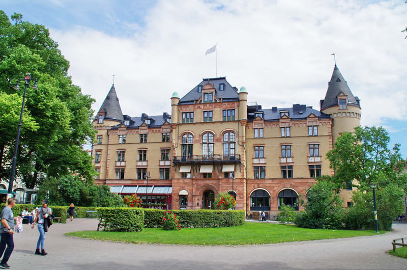Bilden visar en stor byggnad i fokus, omgiven av grönska och ett parkliknande område en solig dag. Byggnaden har en gulbrun fasad med flera fönster och mörka tak, samt två torn med spetsiga tak. En vit flagga är hissad på taket. Framför byggnaden fin