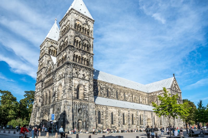 Bilden visar en stor byggnad, en katedral eller kyrka, byggd i sten med två torn i förgrunden och ett långt skepp. Byggnaden har detaljerade tegelfasader och fönster i romansk stil. Taket är grått och har en spetsig del i bakgrunden. Framför byggnade