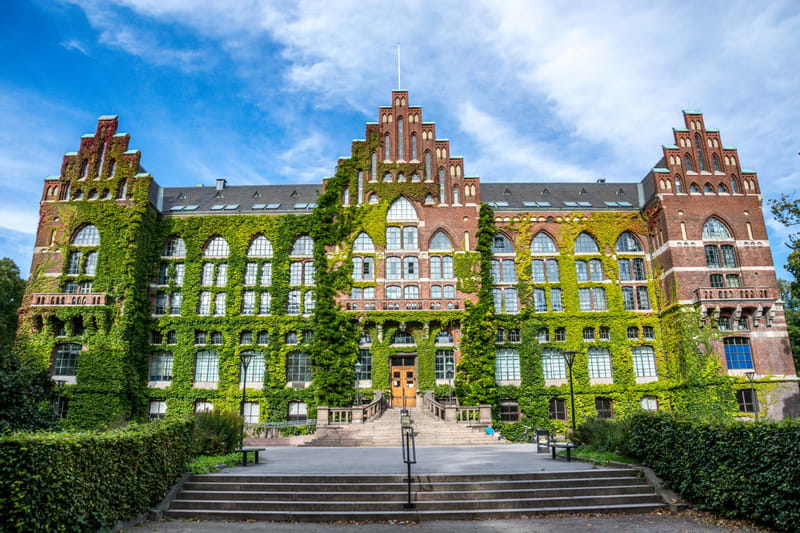 En stor, tegelröd byggnad delvis täckt av grön murgröna syns under en blå himmel med vita moln. Byggnaden har många fönster i olika storlekar och former, samt flera gotiskinspirerade torn och arkitektoniska detaljer. En mittdel med en flaggstång på t