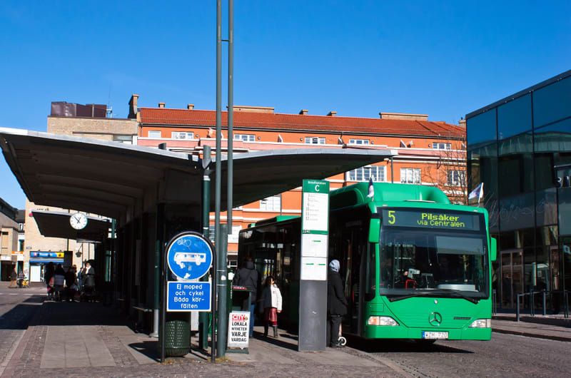 Bilden visar en busshållplats under en klarblå himmel. En grön buss med texten "5 Pilsäker via Centralen" står vid hållplatsen och passagerare verkar vara på väg att stiga ombord eller av. Vid hållplatsen finns det en tidtabell för buss "C" som lista