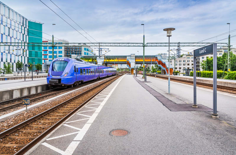 Bilden visar ett tåg på en järnvägsstation under dagtid med en himmel som har moln. Ett blått tåg är synligt på rälsen till vänster i bilden, sedd från plattformen. Texten "Pågatågen" syns på tågets sida. Till höger om tåget löper spåren parallellt m
