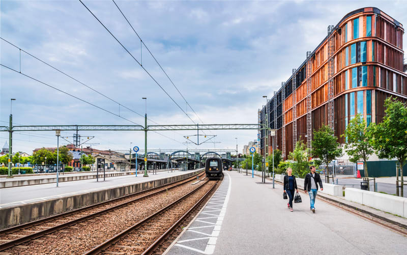 Bilden visar ett tåg som närmar sig en tågstation med flera plattformar och spår. Till höger i bilden finns en stor byggnad med kopparfärgade paneler och fönster som sträcker sig över flera våningar. Framför byggnaden finns träd och på gångvägen går