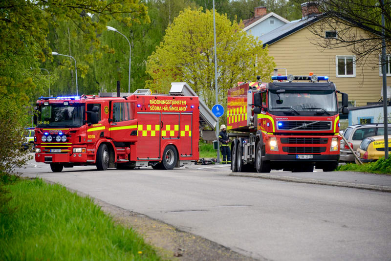 Bilden visar två röda utryckningsfordon, sannolikt brandbilar, parkerade på en asfalterad väg. Fordonen har blåljus som indikerar att de är i tjänst. På sidan av det vänstra fordonet syns texten 'RÄDDNINGSTJÄNSTEN SÖDRA HÄLSINGLAND'. Ett av fordonen