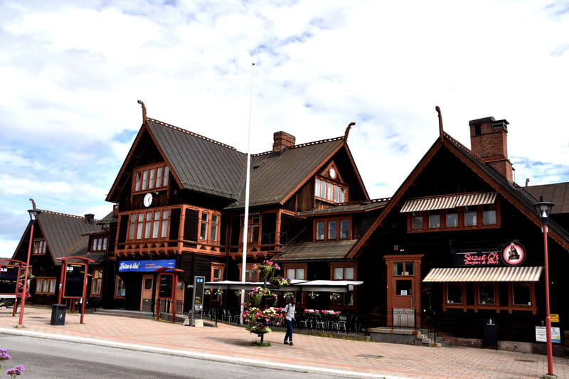 Bilden visar fasaden på en större byggnad med traditionell arkitektur i trä. Byggnaden har mörkt trä och ljusare bruna detaljer, samt tak i mörk plåt eller skiffer. Flera fönsterpartier syns, varav en har en stor klocka ovanpå. Framför byggnaden finn