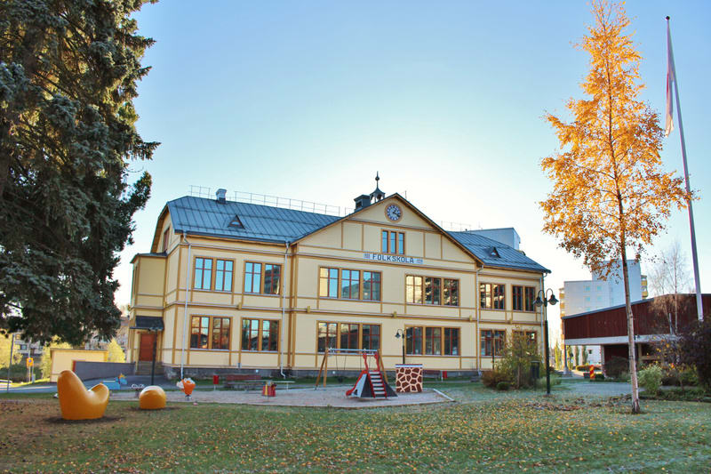 Bilden visar en gulbeige byggnad med mörkgrått tak och många fönster. Ovanför huvudentrén finns texten "FOLKSKOLA" och en klocka. Framför byggnaden finns en lekplats med sandlåda, gungor, en röd rutschkana och en gul klätterställning. På den gröna gr