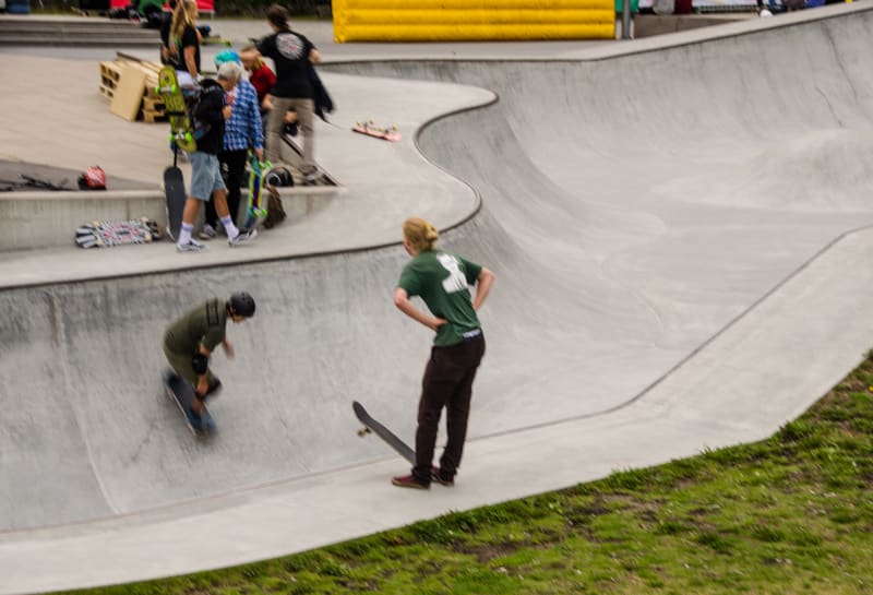Bilden visar en skateboardpark med flera personer. I förgrunden till vänster åker en person skateboard i själva poolen, iklädd en grön tröja och hjälm. Bredvid poolen står en annan person, vänd med ryggen mot kameran, iklädd en grön t-shirt och mörka