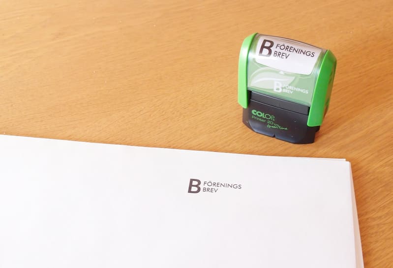 Bilden visar ett skrivbord eller en yta av ljust trä med ett staplat papper i nedre vänstra delen av bilden och en stämpel i övre högra delen. Pappersarken är vita och på det översta arket finns texten "B FÖRENINGS BREV" tryckt i svart, placerad i den övre vänstra tredjedelen. Stämpeln är grön och svart med vita inslag och har texten "B FÖRENINGS BREV" synlig på locket. Dessutom syns texten "COLOP Printer 20 Green Line" på stämpelns svarta del. Bakgrunden är oskarp men har ett mönster av träfibrer. Ljuset kommer från en okänd källa och skapar en mjuk skugga under stämpeln.