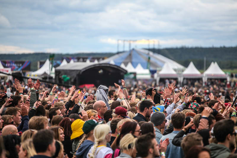 Bilden visar en stor folkmassa vid ett utomhusevenemang. Många människor har sina händer sträckta uppåt, vilket antyder applåder, jubel eller engagemang i evenemanget. I bakgrunden syns tält, en scen eller plattform, samt delar av en större konstrukt