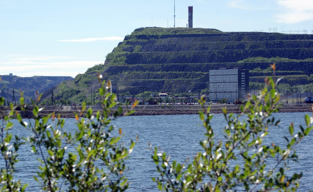 Bilden visar ett landskap med en stor, terrasserad kulle täckt med vegetation i bakgrunden. Ovanpå kullen står en röd- och vitfärgad byggnad och en antenn. Framför kullen, närmare marknivån, syns en stor modern svart byggnad med många fönster. Mellan