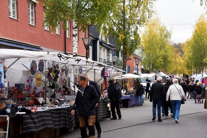 Bilden visar en utomhusmarknad längs en gata med många besökare. I förgrunden till vänster syns marknadsstånd med vita takpresenningar fyllda med diverse varor, inklusive leksaker, smycken och retro-skyltar. Flera personer, både handlare och kunder,