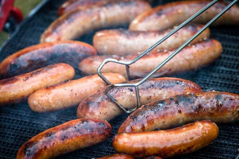 Bilden visar ett antal korvar som grillas på ett grillgaller. Korvarna har olika nyanser av brunt, vissa är mer gyllenbruna medan andra är mörkare och har brända partier. Lätt rök eller ånga stiger från korvarna. En grilltång av metall, med en böjd f