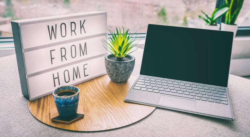 En bild interiört som visar en arbetsplats med en ljuslåda med texten "WORK FROM HOME". Dessutom syns en uppslagen laptop med ett grått tangentbord och en svart skärm. Mellan ljuslådan och laptopen står en krukväxt i en grå kruka. På bordsskivan, int