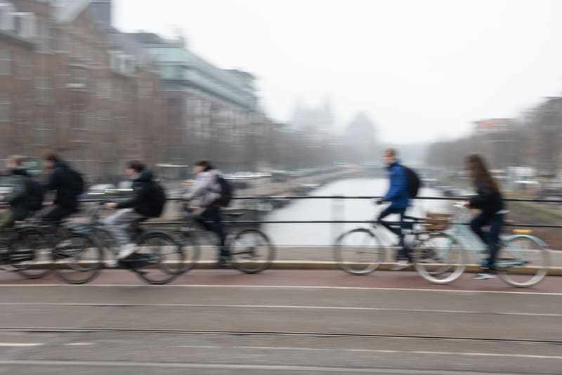 Bilden visar flera personer som cyklar över en bro. Föremålen och personerna i förgrunden är i rörelseoskärpa, vilket indikerar att de rör sig snabbt i sidled, möjligen framför stillastående bakgrund eller att fotografen har panoreringsteknik. I bakgrunden syns urbana byggnader och en kanal som sträcker sig under bron. Himmel är ljus och mulen, vilket skapar en diffus belysning. Cyklisterna bär vardagliga kläder, varav några bär ryggsäckar. Vägen verkar ha en avgränsad cykelbana i rött. Byggnaderna i bakgrunden är suddiga, men verkar vara flerfamiljshus längs kanalen.
