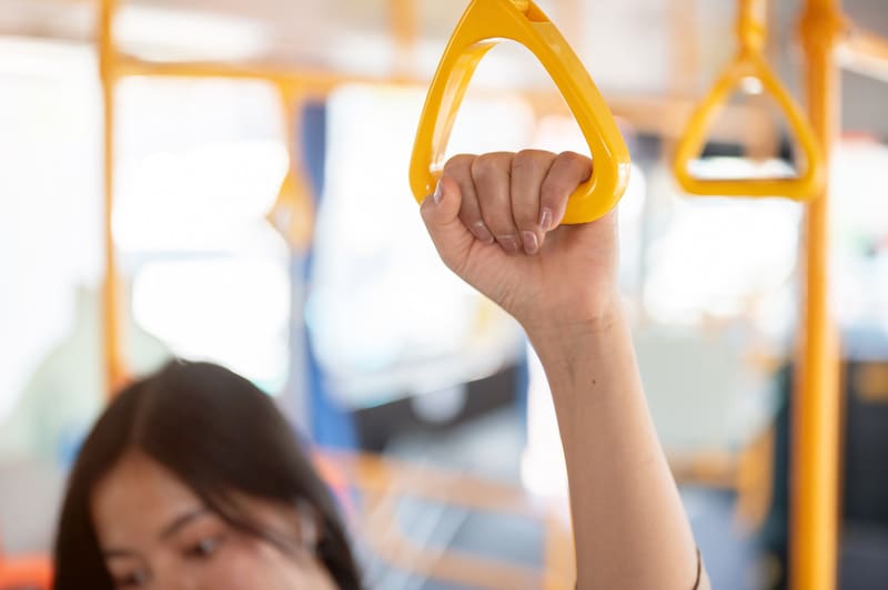 En bild tagen inifrån ett fordon, troligtvis en buss, där en person håller i en gul hållhandtag. Personens arm och hand syns tydligt, med fokus på handen som griper tag om handtaget. Personen har mörkbrunt eller svart hår som syns nedtill i bilden oc