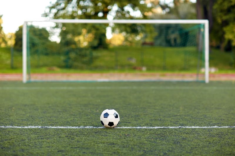Bilden visar en svart och vit fotboll placerad på en konstgräsplan. Fotbollen ligger på en vit linje som sträcker sig horisontellt över bildens nedre del. I bakgrunden, bakom fotbollen, finns ett fotbollsmål med vitt ramverk och ett grönt nät. Bakom målet syns grönskande träd och gräs, vilket indikerar att bilden är tagen utomhus under dagtid. Fokus ligger på fotbollen och den närmaste delen av planen, medan bakgrunden är suddig.