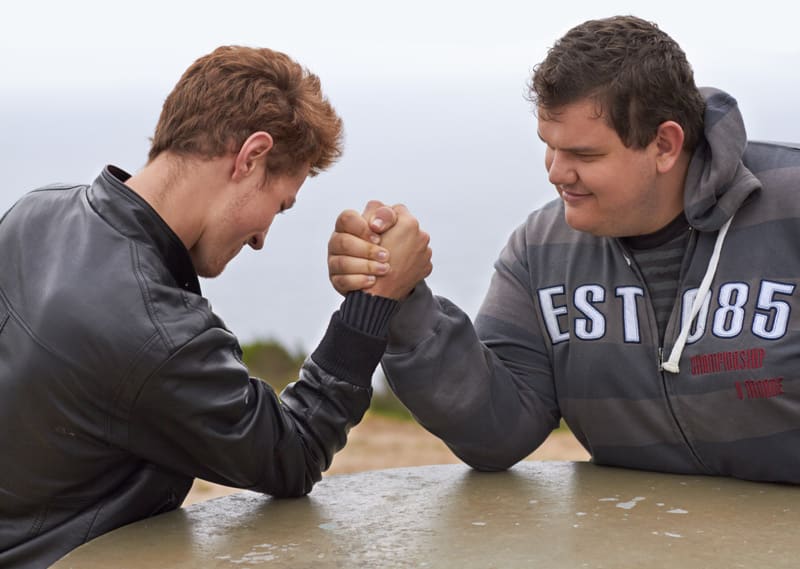 Två män är avbildade utomhus engagerade i en handikappstävling. De står mot varandra med sina högra armbågar placerade på en ljusbrun bordsskiva, hållandes varandras högra händer i ett fast grepp. Mannen till vänster bär en svart läderjacka med elast