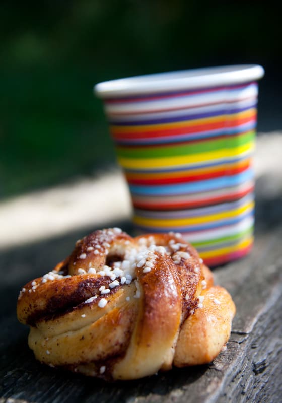 Bilden visar en bulliknande bakelse, möjligen en kanelbulle, toppad med pärlsocker. Bakelsen ligger på en träyta. I bakgrunden syns en färgglad pappmugg med horisontella ränder i olika färger. Bakgrunden är oskarp med gröna inslag, vilket kan tyda på