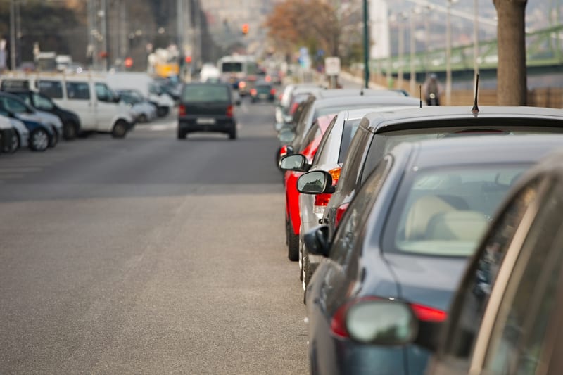 Bilden visar en gata med en rad parkerade bilar längs ena sidan och en väg för trafik i mitten. I förgrunden syns bakdelarna av flera bilar, med en mörkblå bil närmast kameran. Bortom de parkerade bilarna, i blicklinjen, syns en bro med gröna detalje