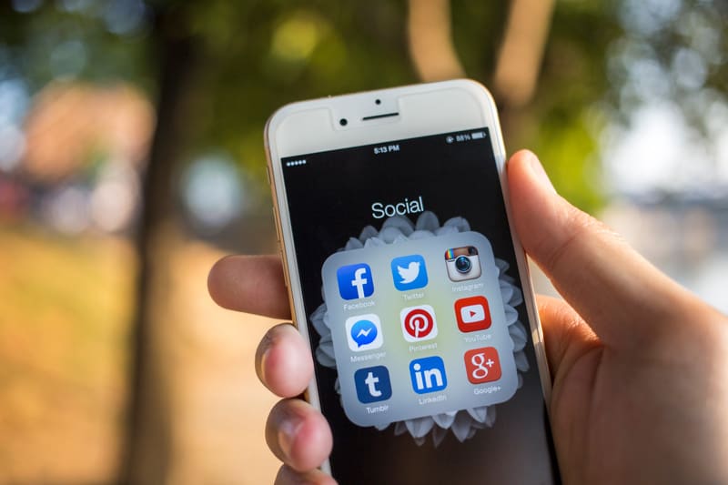 En hand håller en vit smartphone utomhus med en oskarpt grön och gul bakgrund. På telefonens skärm visas en mapp med namnet "Social" som innehåller ikoner för olika sociala medier. Bland ikonerna finns logotyper för Facebook, Twitter, Instagram, Messenger, Pinterest, YouTube, Tumblr, LinkedIn och Google+. Överst på skärmen syns klockan "5:13 PM" och batteristatus "88%". Telefonen är i vertikalt läge och hålls av en person vars fingrar syns runt enhetens kanter.