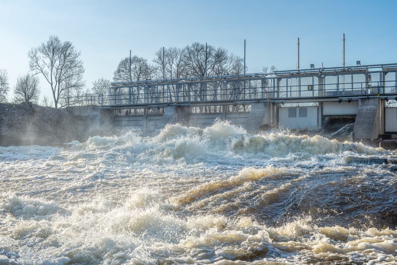 Bilden visar ett kraftigt forsande vattenflöde under en dammkonstruktion. Vattnet skummar och kastar sig fram med stora vågor i förgrunden. I bakgrunden syns en betongdamm med en metallbro över dammluckorna. På bron finns ett metallräcke. Bakom dammen och bron syns kala trädgrenar mot en ljusblå himmel. Delar av dammkonstruktionen är vita med fönster eller ventilationsöppningar. Vattenånga eller dis bildas där vattnet kraschar mot hindren.