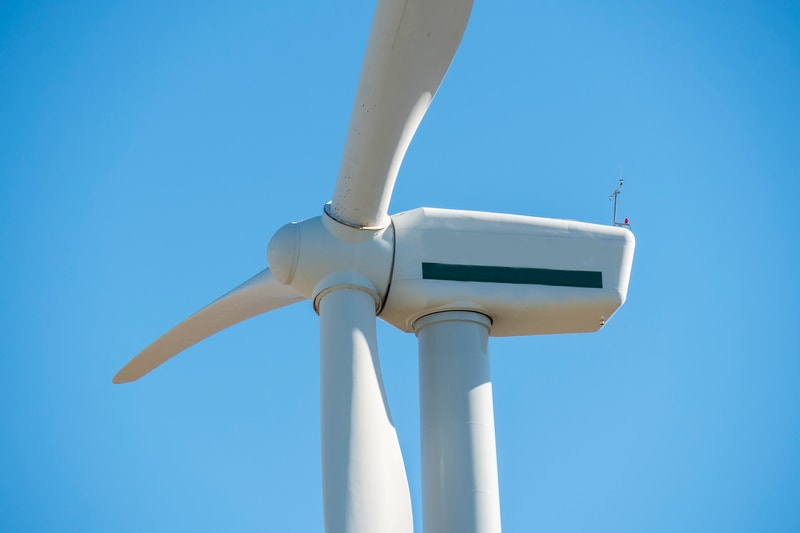 En närbild av den övre delen av ett vindkraftverk med en klarblå himmel i bakgrunden. Två av de vita rotorbladen är synliga, varav ett sträcker sig horisontellt och det andra pekar snett nedåt. Navet och maskinhuset är också vita, med en mörkgrön hor