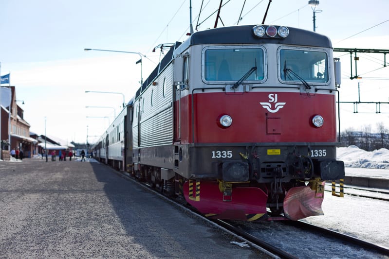 Bilden visar ett rött och grått tåg av modell SJ, nummer 1335. Tåget står på en räls med en plattform bredvid. På framsidan av tåget syns ett snöplogsliknande tillägg. I bakgrunden syns en byggnad som liknar en station eller en perrong. Flera människ