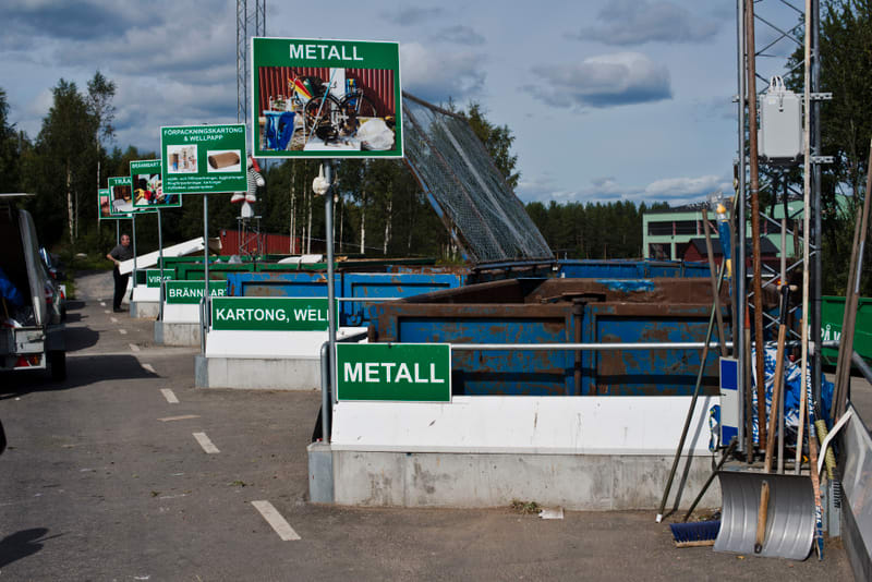 Bilden visar en återvinningscentral en solig dag. I förgrunden syns asfalt med vita streck som avgränsar körbanor. Flera stora blå containrar, delvis rostiga, är placerade i rader och avsedda för olika typer av avfall. Ovanför containrarna finns grön